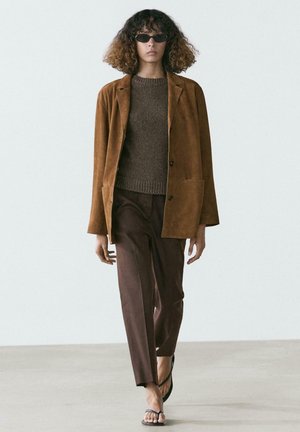 Vrouw met krullend haar die een zwarte zonnebril, een bruine suède jas, een gebreide trui, wijde broekspijpen en zwarte sandalen draagt en naar voren loopt.