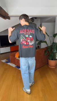 Person innendørs som peker med tomlene mot baksiden av en svart Adidas-skjorte med rød chilipeppergrafikk og teksten "Extra Hot", iført lyseblå jeans.