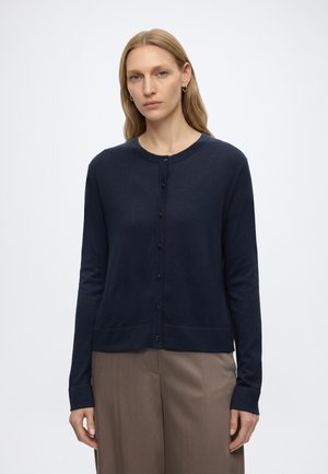 Femme portant un cardigan bleu marine boutonné et un pantalon marron, debout devant un fond blanc uni, regardant devant elle.