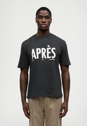 Homem a usar uma t-shirt preta com o texto "APRÈS" e uma pequena impressão, combinado com calças castanhas texturizadas, de pé contra um fundo branco liso.