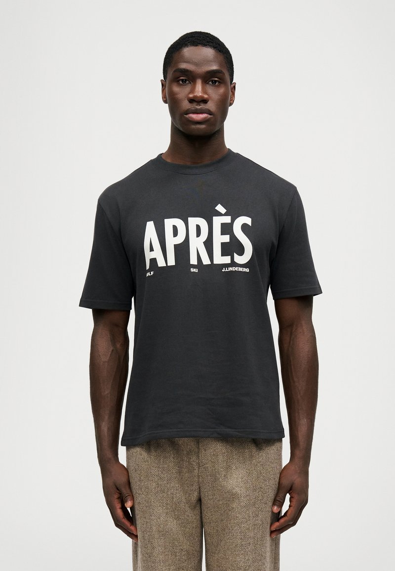 Άνδρας φορώντας μαύρη μπλούζα με κείμενο "APRÈS" και μικρή εκτύπωση, σε συνδυασμό με καφέ υφασμάτινο παντελόνι, που στέκεται μπροστά σε λευκό φόντο.