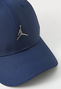 Mørkeblå cap lavet af tekstureret stof, med et sølvfarvet Jumpman-logo på fronten, buet skygge og forstærkede syninger.