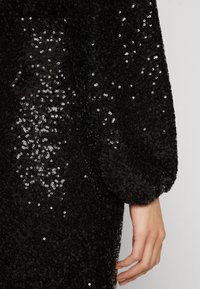 Svart paljettklänning med detaljer som har en texturerad yta med reflekterande paljetter. En samlad, voluminös ärm syns tillsammans med en hand.