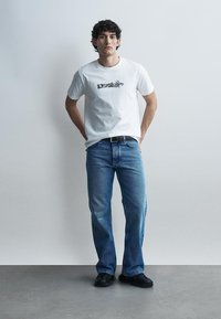 Giovane uomo con capelli ricci e scuri che indossa una t-shirt bianca con grafica, jeans blu e scarpe nere, in piedi davanti a uno sfondo chiaro e uniforme.