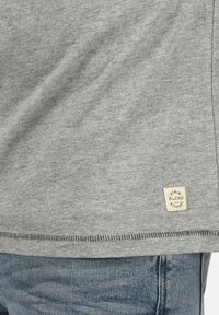 Pelēka kokvilnas T-krekls ar redzamu šuvumu apakšmalā, ar kvadrātveida marķējumu, kas norāda "DENIM BLEND with ATTITUDE" bežā un melnā krāsā.