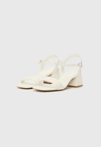 White heeled sandals with wide front straps and a block heel. The straps have a smooth finish, and an adjustable ankle buckle is featured.