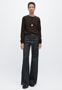 Brun tröja, mörka jeans med vida ben och svarta kraftiga skor. Silverhalsband med hänge, enkel textur, neutrala färger, avslappnad stil.