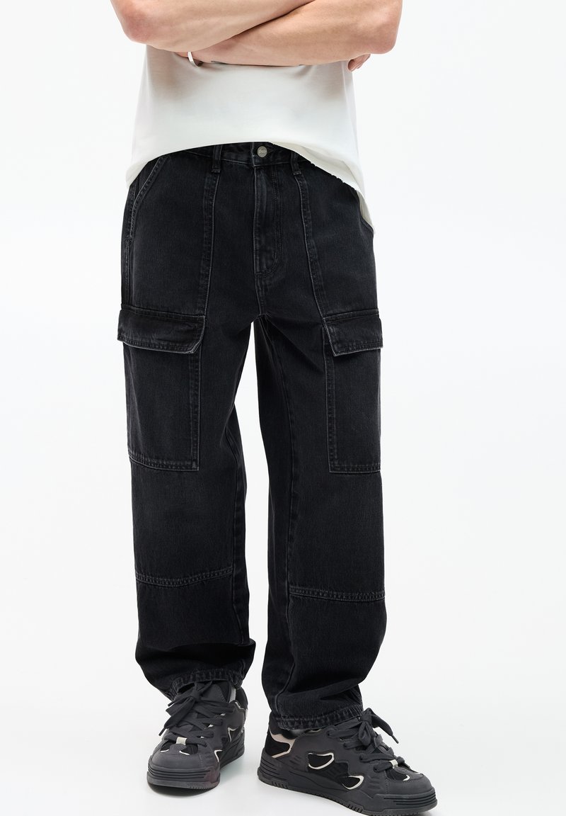 Schwarze Denim-Cargohosen mit mehreren Taschen, lockerer Passform, sichtbaren Nähten und elastischen Bündchen. Getragen mit grauen Sneakern.