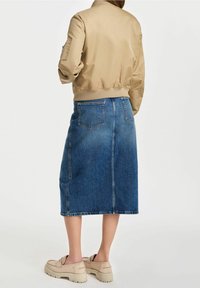 Chaqueta bomber beige con cinturilla de canalé, combinada con una falda midi de mezclilla azul clara que presenta una costura central y bolsillos traseros. Zapatos beiges de tipo slip-on.