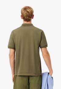 Uomo con i capelli biondi corti che indossa una polo verde oliva e pantaloni corti, tiene una giacca blu chiaro nella mano destra, di spalle.
