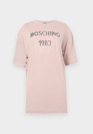 Nő állva, világos rózsaszín oversized Moschino 1983 pólót, laza világoskék farmert és fekete, nyitott orrú magassarkú szandált viselve, fehér háttér előtt.
