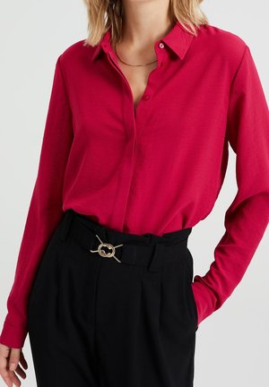 Donna che indossa una camicia rossa a maniche lunghe con bottoni infilata in pantaloni neri a vita alta con cintura con fibbia dorata e una mano in tasca.