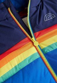 Színes gyermekdzseki, amelynek tengerészkék alapszíne van, vízszintes szivárványcsíkokkal és sárga cipzárral, kör alakú húzózsinórral.