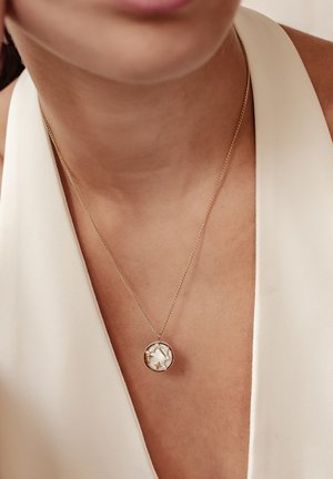 Goldene Halskette mit einem runden Anhänger, der ein weißes Steindesign und kleine Akzente aufweist. Die Kette ist fein und zart.