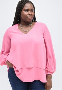 Γυναίκα με μακριά πλεξούδα, φορεμένη με ροζ μπλούζα μακρυμάνικη και μπλε τζιν, στέκεται με το ένα χέρι στην τσέπη, κοιτάζοντας προς τα πλάγια.