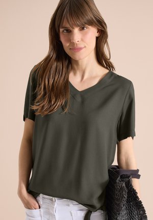 Kobieta z długimi brązowymi włosami, ubrana w oliwkowo zielony top z krótkim rękawem i dekoltem w serek, białe spodnie, trzyma czarną plecioną torbę, jedną ręką w kieszeni.