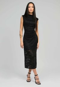 Robe midi en dentelle noire avec une silhouette ajustée, un col montant, des manches courtes et un panneau inférieur transparent. Des talons noirs à lanières accentuent le look.