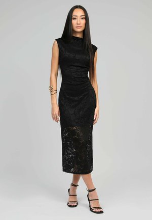 Robe midi en dentelle noire avec une silhouette ajustée, un col montant, des manches courtes et un panneau inférieur transparent. Des talons noirs à lanières accentuent le look.
