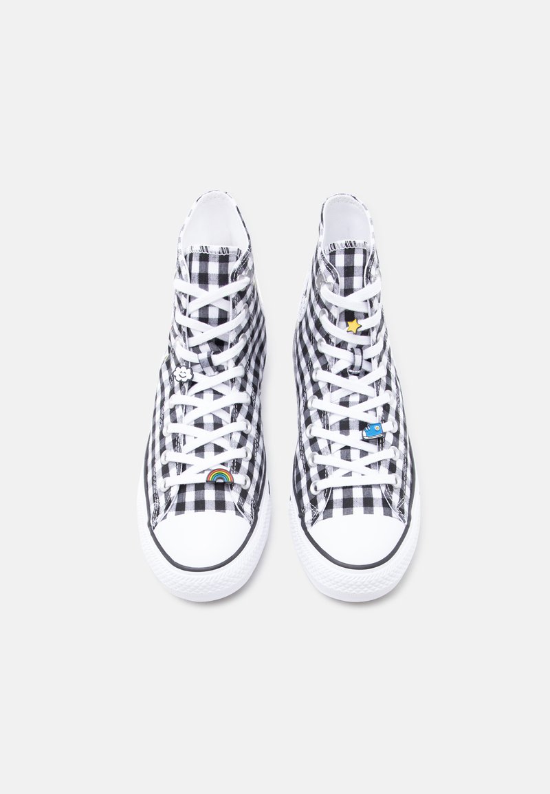 Sneakers alte a quadretti bianchi e neri con lacci bianchi e piccoli ciondoli colorati sui lacci, su uno sfondo bianco semplice.