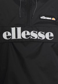Schwarze Windjacke mit einem Viertel-Reißverschluss, mit großem weißen "ellesse"-Logo und kleinem ellesse-Wappen auf der Brusttasche. Glatte Textur.