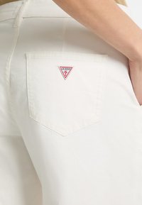 Balti džinsai su trikampiu raudonu ir baltu "GUESS?" logotipu ant galinio kišenės, su kišenės dizainu ir lygiu paviršiumi.