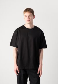 Camiseta negra de algodón con mangas cortas y escote redondo. Presenta un borde contrastante en blanco a lo largo del cuello y las mangas.