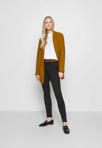 Hořčicově zbarvený blazer, bílý tričko, černé skinny džíny a černé mokasíny. Má hladkou tkaninu a strukturovaný design. Hnědý pásek jako akcent.