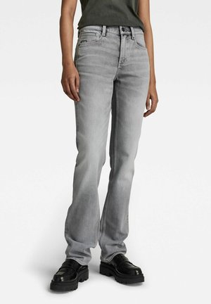 Henkilö, joka pukeutunut vaaleanharmaisiin suoriin farkkuihin ja mustiin tukeviin loafereihin, seisoo yksinkertaisen valkoisen taustan edessä.