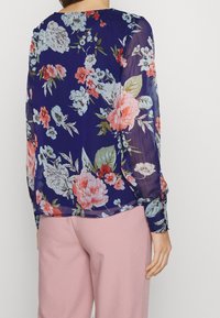 Blouse fleurie en tissu bleu transparent, avec de grandes fleurs roses et bleu clair. Les poignets des manches présentent des accents floraux. Pantalon rose clair.