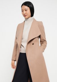Frau in einem beigen langen Mantel über einem hellen Rollkragenpullover und dunklen Hosen, die mit einer Hand in der Manteltasche vor einem weißen Hintergrund steht.
