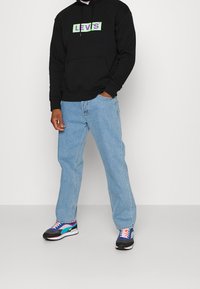 Sweat à capuche en coton noir avec un logo vert et violet, jean bleu clair à jambe droite, et baskets multicolores avec des accents noirs et blancs.