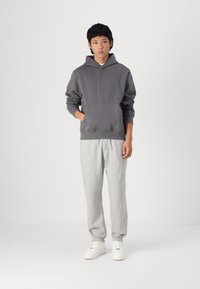 Grå hettegenser med frontlomme, kombinert med lys grå joggebukser og hvite sneakers, står foran en ensfarget bakgrunn.
