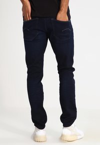 Vaqueros de mezclilla azul oscuro con un corte ajustado, que cuentan con dos bolsillos traseros y detalles de costura sutiles. Combinados con zapatillas de color claro.