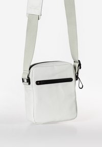 Borsa a tracolla bianca realizzata in materiale liscio, con una tasca frontale con zip, tracolla regolabile e dettagli neri. Design rettangolare compatto.