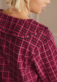 Sarkans un melns rūtainā blūze ar viļņotu apkakli un teksturētu audumu, demonstrējot režģa rakstu un plecu detalizāciju ar pintēm.