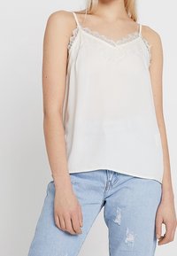 Bílá saténová košilka se krajkovým lemováním na výstřihu a ramínkách. V páru s světle modrými roztrhanými džíny. Jednoduchý, elegantní design.