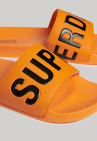 Oranje schuifsandalen met een gestructureerde rubberen zool en een brede band met het woord "SUPER" in vette zwarte letters.
