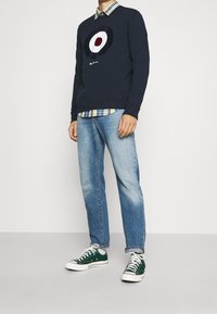 Granatowa bluza z okrągłą grafiką, noszona na koszuli w kratę. Do tego jasnoniebieskie dżinsy i zielone trampki z białymi podeszwami.