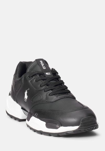 Svarte idrettssko laget av syntetisk skinn, med en teksturert overdel, polstret krage, gummisåle og hvite detaljer. Nøkkel-logoen er vist.