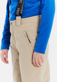 Beige skibroek met een tailleband en verstelbare bretels, voorzien van een ritszak aan de zijkant en gestikte naden, gedragen met een blauwe fleecetop.
