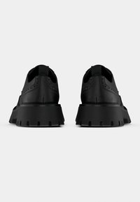 Μαύρα αθλητικά παπούτσια με υφασμένο δερμάτινο επάνω μέρος, επενδεδυμένο γιακά και παχιά, χοντρή λαστιχένια σόλα με βαθιές αυλακώσεις. Χωρίς ορατά κορδόνια.