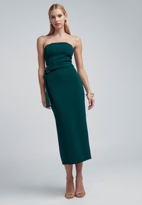 Abito midi color teal senza spalline con corpetto aderente e cintura in vita. Presenta tessuto strutturato e spacco laterale. Indossato con tacchi trasparenti.