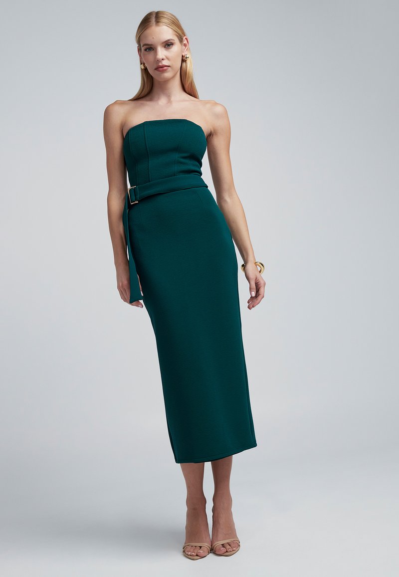 Abito midi color teal senza spalline con corpetto aderente e cintura in vita. Presenta tessuto strutturato e spacco laterale. Indossato con tacchi trasparenti.
