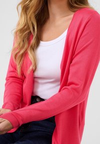 Roze cardigan van zacht materiaal, met een ontspannen pasvorm en lange mouwen, gecombineerd met een witte top. De achtergrond is eenvoudig en neutraal.