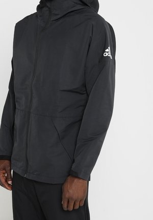 Giacca nera con cappuccio Adidas con zip frontale, tasche laterali e logo Adidas sulla parte superiore della manica sinistra indossata da una persona in piedi.