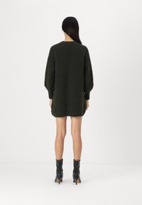 Abito lavorato a maglia verde scuro con un taglio morbido, texture a coste e maniche lunghe, indossato con stivali neri alla caviglia. La lunghezza arriva a metà coscia.