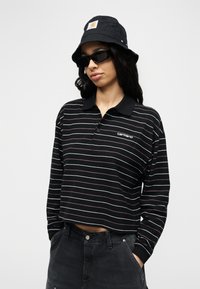 Γυναίκα που φοράει μαύρο καπέλο τύπου bucket, μαύρα γυαλιά ηλίου, ριγέ μαύρο cropped μακρυμάνικο πόλο πουκάμισο και σκούρο παντελόνι με ψηλή μέση, με τα χέρια στις τσέπες.