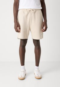 Shorts texturés beige avec une taille élastique et un cordon de serrage. Présentent des poches latérales et une marque subtile au niveau de l'ourlet. Associés à des baskets blanches.