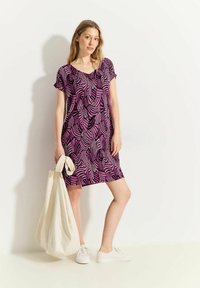 Lila Kleid mit einem Wellenmuster in Pink, kurze Ärmel, lockere Passform, knie-länglich, kombiniert mit einer weißen Tragetasche und weißen Turnschuhen.
