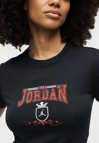 Γυναίκα με σγουρά μαλλιά που φοράει μαύρο t-shirt με κόκκινο κείμενο «1984 JORDAN» και λευκό λογότυπο Jumpman μέσα σε ασπίδα με στέμμα.